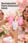 Vertical bachelorette party photo with three hands raising small bridal-themed cups for a toast: two pink cups show a ring-finger illustration and “she said yes!” lettering, while a white cup reads “i said yes!” in pastel lettering. Two women in pink heart-shaped sunglasses smile in the background beside pink-and-white balloons, flowers, candles, macarons on a gold tray, and a marble tabletop. The headline at the upper left reads “Bachelorette Party Drinkware Ideas.”