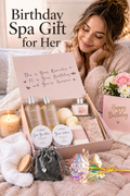 A blush pink birthday spa gift box arranged on soft neutral bedding while a woman in a plush robe smiles behind it. The open box shows LOVERY bubble bath, shower gel, body scrub, body lotion, bath salts, candle and bath items. A heart necklace in a dark pouch, iridescent decorative flower, white fuzzy socks, fresh pink and white flowers, and a Happy Birthday card surround the box. Headline reads “Birthday Spa Gift for Her.”