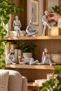 Vertical lifestyle photo of a warm wood bookshelf in a softly lit living room, styled with leafy plants, neutral books, ceramics, framed art, a lamp, and six white reading-woman figurines covered in black letter print. Five figures sit or recline across the shelves holding rust, burgundy, or red books, while a cream-sleeved hand places a sixth seated figure on the upper shelf. Natural sunlight, a textured chair, and a candle complete the cozy reading-corner scene.