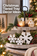 Vertical Christmas lifestyle photo of a distressed white two-tier tray on a rustic tabletop, styled with evergreen branches, red berries, pinecones, a glowing glass candle, a snowy bottle-brush tree, and a small lighted cream house. Three white wooden freestanding snowflakes with visible natural wood edges are arranged across the lower tier. Warm tree lights blur in the background, and the headline “Christmas Tiered Tray Decor Ideas” appears in white at the upper left.