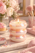 A pink iridescent bubble-glass scented candle burns at the center of a feminine vanity scene on a cream marble surface. The rounded three-tier vessel reflects soft peach, gold, and rainbow tones around pale wax and a single visible flame. Pink and white flowers fill the upper left, while a frosted cupcake, a bowl of colorful sprinkles, makeup brushes, jewelry, blush-toned boxes, and a plush pink textile create a warm, softly lit decorative setting.
