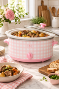 Vertical lifestyle image of a pink-and-white gingham slow cooker centered on a light stone kitchen counter. The cooker is filled with a hearty beef stew with potatoes, carrots, and herbs, while its clear glass lid rests partly open across the top. A plated serving of stew sits on a matching gingham cloth in the foreground, with sliced bread, chopped herbs, flowers, wooden boards, utensils, greenery, and soft daylight from a nearby window completing the warm kitchen scene.
