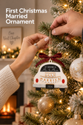 Vertical holiday lifestyle photo of two hands hanging a newlywed Christmas ornament on a warmly lit tree. The ornament is shaped like the rear of a white car with red taillights, silver bumper, black wheels, a black window reading “1ST CHRISTMAS AS Mr. & Mrs.,” a layered “JUST MARRIED” banner, black “2026” plate, and a red-and-black plaid bow with wooden beads. Warm tree lights and a fireplace glow behind it. Headline at upper left reads “First Christmas Married Ornament.”