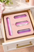 Vertical lifestyle image of a blush-pink food storage bag organizer fitted inside an open light-wood kitchen drawer beneath a pale marble countertop. The organizer has four dispensing openings with clear zip-top bags visible, including pink and purple zipper seals, plus a small PH mark on the front. Brass drawer hardware, a pink-striped cloth, a pink planter with white flowers, and soft natural sunlight create a polished pastel kitchen scene with warm shadows.