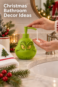 Vertical Christmas bathroom scene with the headline “Christmas Bathroom Decor Ideas” at the upper left. A bright green Grinch-face pump dispenser sits on a light marble vanity beside a white sink while two hands press the pump and catch white soap. Evergreen branches, red berries, warm fairy lights, a glass candle holder, a ceramic cup, a tree-embroidered towel, and a round wood-framed mirror create a cozy holiday setting that shows the dispenser in use.