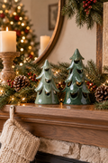 Two glossy green ceramic Christmas tree figurines, one light sage and one deeper forest green, sit side by side on a wooden mantel. Evergreen garland, pinecones, red berries, warm string lights, pillar candles, a round framed mirror, a softly blurred Christmas tree, and a cream knit stocking create a cozy holiday setting. The figurines have layered branch shapes and dark decorative cutouts, while warm ambient lighting keeps both trees clearly visible as a festive focal point.