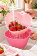 A bright lifestyle kitchen scene shows a pale pink heart-shaped colander held under a running brass-toned faucet, filled with fresh red strawberries as water rinses over the fruit and drains through the perforations. A deeper pink matching bowl sits directly below to catch the water. Hands with pale pink nails hold both pieces over a white sink, with marble counters, pink flowers, greenery, a utensil crock, and a striped pink towel softly blurred in the background.