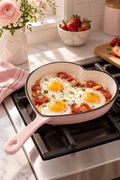 Vertical lifestyle photo of a pale pink heart-shaped skillet with a cream enamel interior resting on a black stovetop grate in a bright kitchen. Three sunny-side-up eggs cook inside with roasted cherry tomato halves, herbs, and browned edges. The long pink handle points toward the lower left. A pink striped towel lies beside the stove, while soft-focus flowers, a bowl of strawberries, sliced berries on a wood board, and a pink jar fill the warm background.