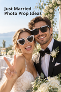 Vertical wedding lifestyle photo with the headline “Just Married Photo Prop Ideas” at the upper left. A smiling newlywed couple poses outdoors beside the water and a white floral wedding arch. The bride wears white heart-shaped sunglasses edged with pearl-like beads and letter beads reading “JUST MARRIED,” while the groom wears black square sunglasses with matching letter beads. White roses and greenery fill the foreground, with a reception table softly blurred behind them.
