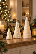 Vertical lifestyle photo of three glowing white ceramic Christmas tree figurines in graduated sizes, each topped with a gold star, arranged on a rustic wooden console. Warm light shines through small round openings in the trees. Evergreen garland, frosted pinecones, red berries, candles, wrapped gifts, and soft golden fairy lights surround the display, with a blurred Christmas tree and gold-framed mirror in the background. The scene feels warm, festive, and illuminated.