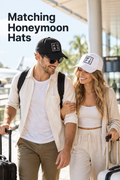 Vertical lifestyle photo of a newlywed couple walking through a bright airport with rolling luggage. The man wears a black baseball cap with a white square patch reading H U over B S, while the woman wears a white cap with a black square patch reading W I over F E. They smile at each other in light travel clothes beside terminal windows, palm trees, and a blurred airplane. Large charcoal headline text at the upper left reads “Matching Honeymoon Hats.”