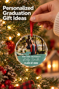 Vertical holiday lifestyle photo of a hand in a cream knit sleeve hanging a round, faceted personalized graduation ornament from a warmly lit Christmas tree. The ornament shows a graduate in a black cap-and-gown outfit above a teal panel reading “NEW YORK UNIVERSITY,” “Kelly Smith,” and “CLASS OF 2026,” with a small graduation-cap graphic. Pine branches, red ornaments, frosted accents, and golden lights fill the scene. Headline: “Personalized Graduation Gift Ideas.”