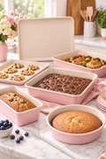 Vertical lifestyle image of a blush-pink and cream bakeware set arranged on a white marble kitchen counter in soft daylight. The square pan holds chocolate brownies, the round pan holds a golden cake, the loaf pan holds a blueberry loaf, a shallow sheet holds chocolate chip cookies, and a larger roasting pan contains roasted chicken with vegetables. A heart-textured cream baking sheet stands behind the pans, with pink flowers, utensils, plates, and a patterned towel nearby.
