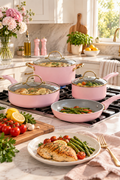 Vertical lifestyle image of a coordinated pastel pink cookware set with gold-tone handles arranged on a black gas cooktop in a bright modern kitchen. A large stockpot and two covered pans hold cooked food under clear glass lids, while an uncovered frying pan contains asparagus and roasted tomatoes. Fresh tomatoes, lemons, garlic, herbs, flowers, a cutting board, and a plated chicken dinner frame the scene on pale marble counters in warm natural sunlight.