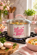 Vertical lifestyle photo of a stainless steel stock pot with pale pink side handles and a matching pink lid handle, centered on a gas stovetop in a bright kitchen. The pot shows the pink heart-filled ‘That’s hot’ graphic on its front, with a clear glass lid resting above steaming soup with pasta, carrots and herbs. Pink bowls, bread, garlic, parsley, grated cheese, flowers and wooden utensils surround the stove, with soft daylight and warm neutral counters in the background.