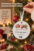Vertical Christmas photo of a hand in a cream knit sleeve hanging a round white pregnancy announcement ornament on a warmly lit evergreen tree. The ornament shows red berries, holly, poinsettias, a baby bottle, bells, and black baby footprints around “MERRY CHRISTMAS,” “Coming soon...,” and “SEE YOU IN 2026.” Red and gold ornaments, pinecones, plaid ribbon, glowing lights, gifts, and a blurred fireplace fill the background. Headline: “Christmas Pregnancy Announcement Ideas.”
