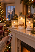 Vertical Christmas lifestyle scene showing three snowman glass flameless pillar candles glowing on a white fireplace mantel in different heights. A hand in a knit sleeve holds the white remote toward the candles. Evergreen garland, red berries, pinecones, ornaments, and warm string lights frame the display, with a softly blurred Christmas tree and snowy window in the background. The candle sleeves show winter cabins, trees, snowflakes, a snowman, and a deer.