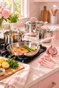 Vertical lifestyle photo of a coordinated stainless steel cookware set with blush pink handles in a bright pastel kitchen. A wide frying pan sits over a gas flame with seared salmon, asparagus and lemon, while a saucepan holds a chunky soup and two lidded pots rest nearby on the marble counter. Pink heart-shaped measuring cups and spoons sit at the right, with lemons, parsley, flowers, a striped towel, cutting boards and pink cabinetry completing the scene.