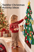 Vertical holiday lifestyle photo showing a toddler in a red Fair Isle sweater and tan pants decorating a large green felt Christmas tree mounted on a cream wall at home. The child holds a Santa ornament while reaching toward colorful felt pieces including snowflakes, candy canes, reindeer, stockings, gifts, and a yellow star topper. A lit Christmas tree and wrapped presents glow in the background. Headline text reads “Toddler Christmas Activity Ideas.”