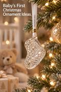 Vertical holiday lifestyle image showing a clear cut-crystal baby boot ornament hanging from a snow-dusted Christmas tree by a white Waterford ribbon and gold 2026 collar. Warm white lights glow around the ornament, with a softly blurred nursery scene behind it featuring a teddy bear, crib rails and a wrapped gift. The top-left headline reads “Baby’s First Christmas Ornament Ideas.” The composition uses warm neutral tones and shallow depth of field.