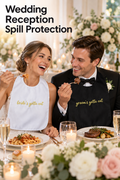 Vertical wedding reception photo of a smiling bride and groom seated together at an elegant dinner table. The bride wears a white adult bib with gold script reading “bride’s gotta eat,” while the groom wears a black bib with matching gold “groom’s gotta eat” embroidery. They hold forks over plated pasta and steak, with candles, champagne glasses, white and blush flowers, and greenery around them. Headline at upper left reads “Wedding Reception Spill Protection.”