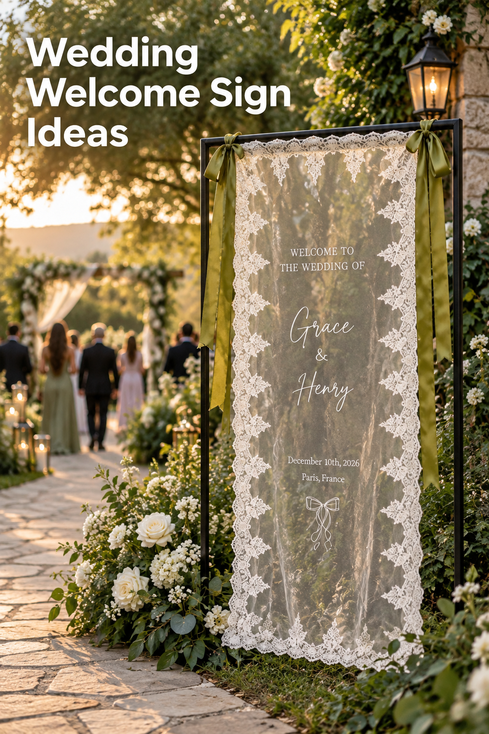 Vertical Pinterest image showing a romantic outdoor wedding entrance at golden hour. A sheer ivory wedding welcome banner with ornate lace edging hangs from a black metal frame and is tied with olive green satin ribbons. White roses, greenery, lanterns, a stone path, and softly blurred wedding guests lead toward a floral ceremony arch. The banner reads “Welcome to the Wedding of Grace & Henry,” and the headline above says “Wedding Welcome Sign Ideas.”