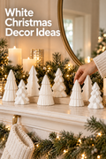 Vertical holiday mantel photo showing eight glossy white ceramic Christmas tree figurines arranged at varied heights on a white mantel among evergreen garland, warm fairy lights, pillar candles, and gold accents. A hand in a cream knit sleeve adjusts one tree near the right side, with a large brass-framed mirror and softly lit Christmas tree in the background. A cream stocking hangs below the mantel. The upper-left headline reads “White Christmas Decor Ideas.”