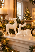 Vertical lifestyle photo of two glossy white reindeer figurines arranged on a cream fireplace mantel for Christmas, with one deer standing and the other resting in front of layered evergreen garland. Warm white string lights, pinecones, gold ornaments, pillar candles, and a gold-framed mirror create an elegant neutral holiday scene. A softly blurred lit Christmas tree glows in the background, while greenery drapes over the mantel edge in the foreground.