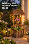 Vertical garden lifestyle image showing a rustic wooden wishing well planter beside a home at dusk. The well has a shingled roof, a small panel mounted near the roofline, warm string lights, a hanging wooden bucket filled with pink flowers, and a large base planter overflowing with mixed blooms and greenery. Stone pavers, shrubs, white flowers, and a small path light surround it. White headline text at the upper left reads “Wishing Well Garden Decor Ideas.”